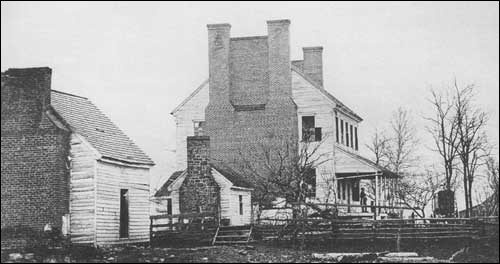 Black and White Photograph of a woman standing on the front porch of a large, double-chimney house separated by a rail fence from a much smaller house.