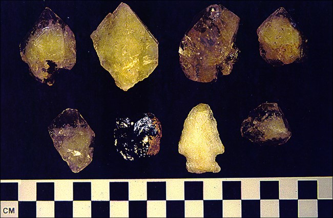 Four dark brown and yellow quartz crystals on top row. Two black and dark gray quartz crystals on bottom left. Next to that is a beige, arrow-shaped, prehistoric quartz point. On far bottom right is a dark brown fragment of galena mineral.