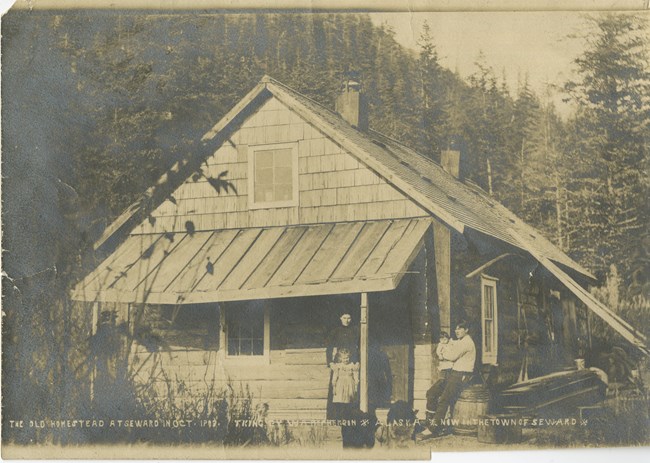 A woman and young girl stand outside the front door of a cabin, while an older man sits nearby holding a child in his arms.