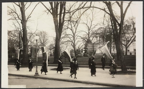 Photograph of suffragists marching in contingent with banners, picketing outside on the sidewalk in front of the White House. A woman skates by in foreground on roller skates, and a man in bowler hat strolls down sidewalk near fence.