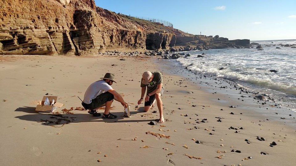Two people collecting sand samples on a beach bellow rocky bluffs