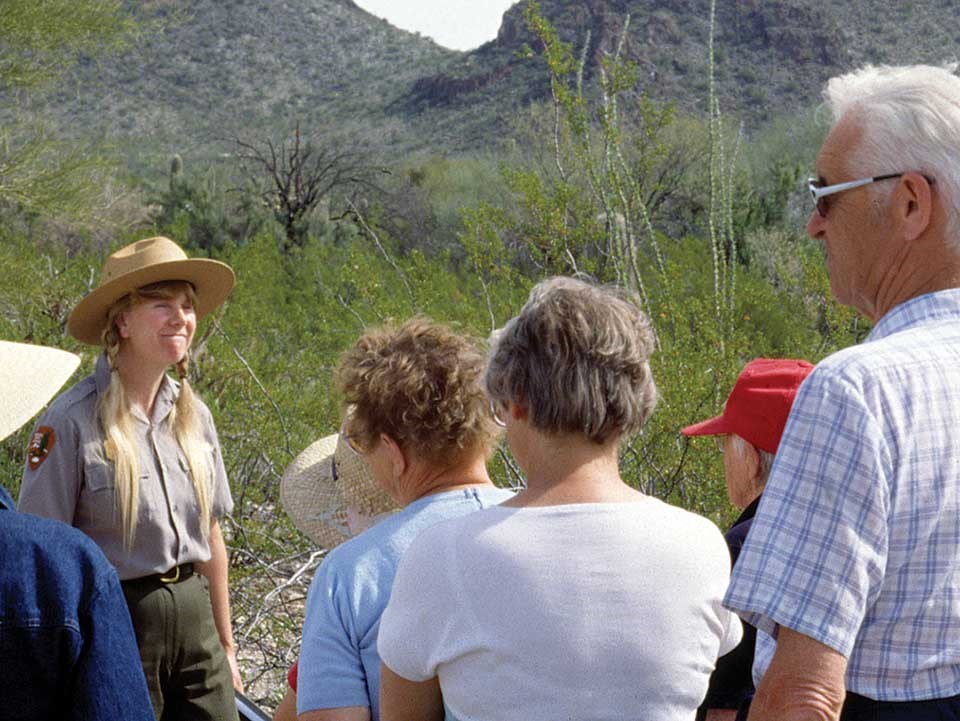A ranger-naturalist conducts an "ant walk" near the park visitor center (1987). A ranger wearing a flat hat smiles at a group of visitors.