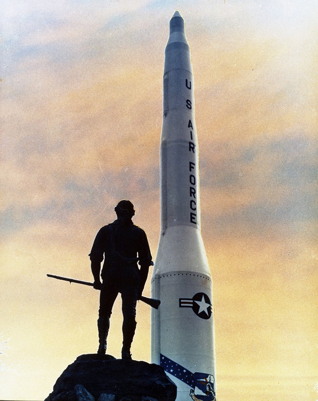 A tall missile model stands behind a revolutionary war soldier statue at sunset