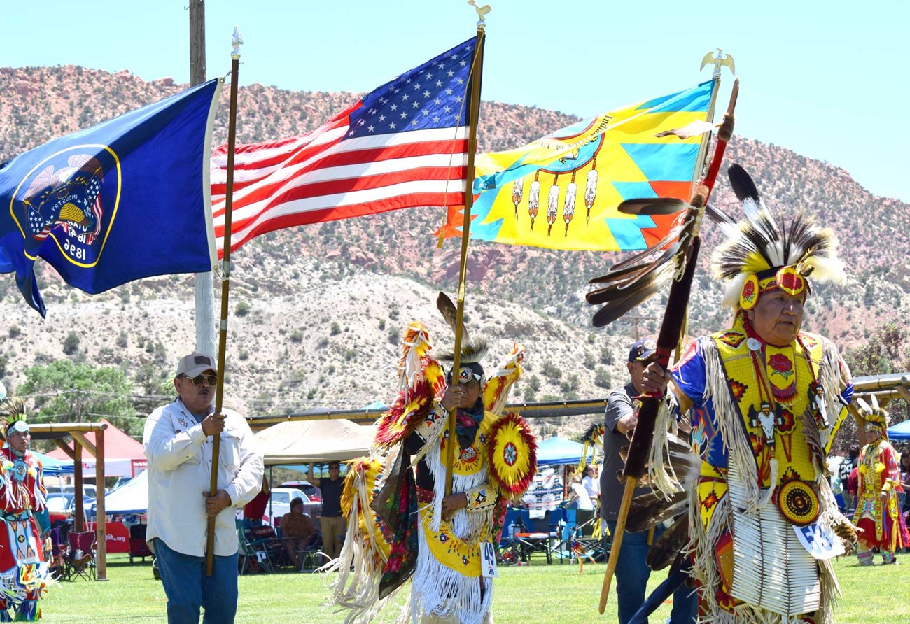 Three Paiute men in ceremonial dress under three flags: American, Utah and Paiute.