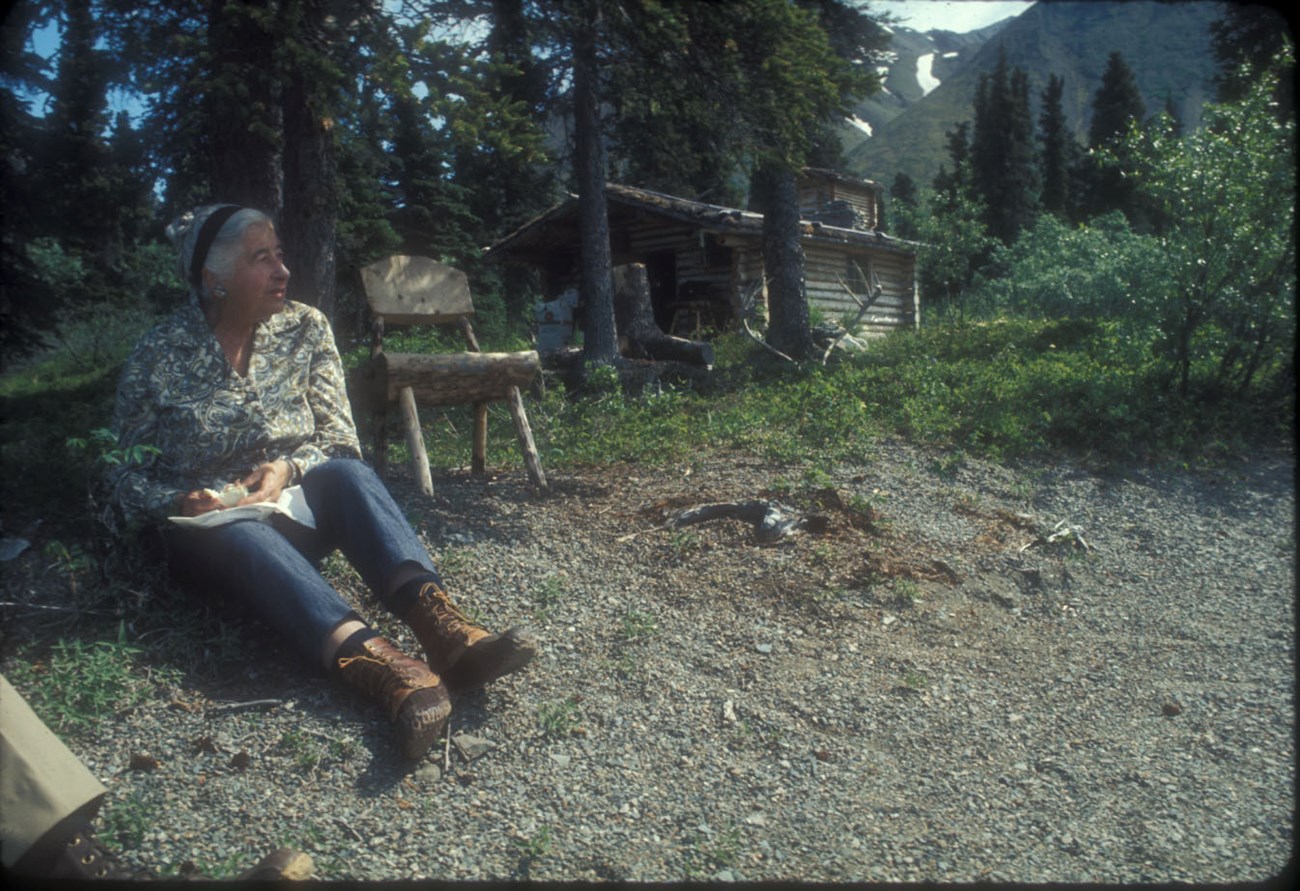 Murie at Richard Proenneke's Cabin Image of woman sitting on a beach with cabin and chair in background.