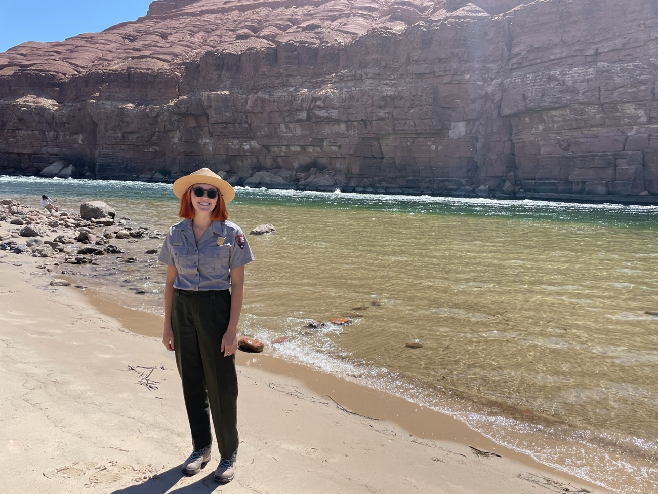 Person in ranger uniform stands near rushing river water's edge. Tall canyon wall in the background.
