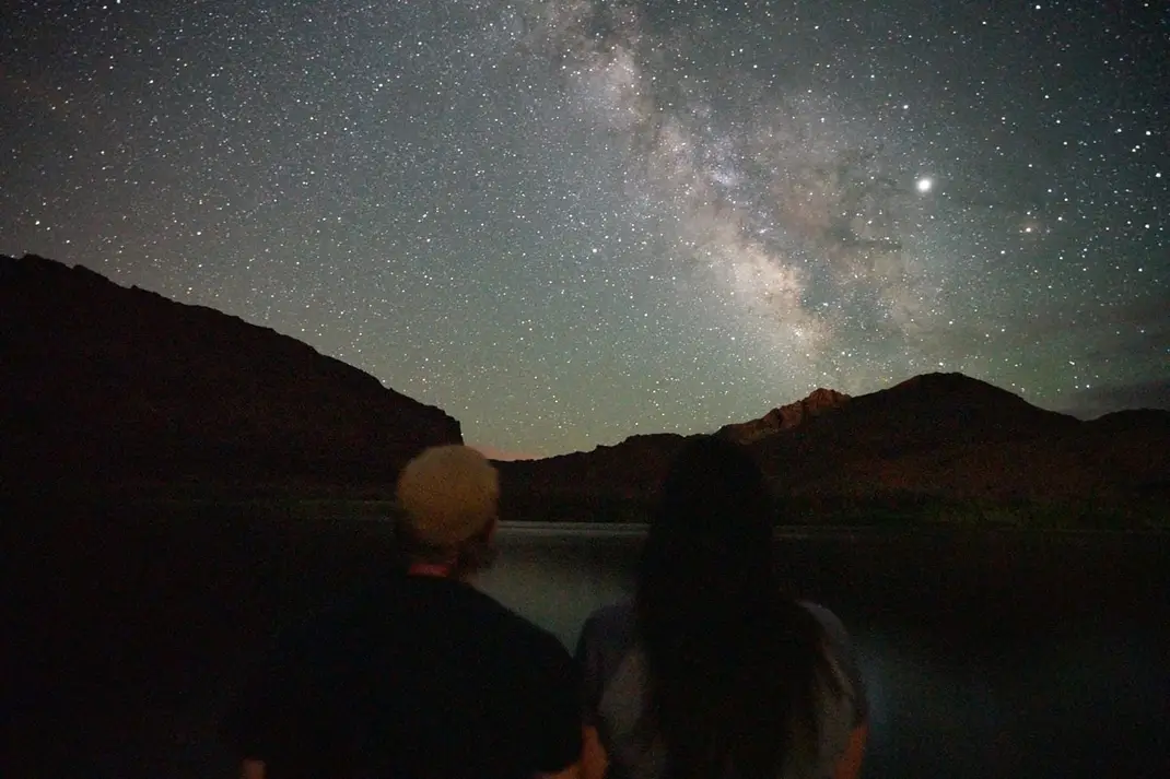 lt= Two people stand in the dark looking up at the Milkey Way in the sky.