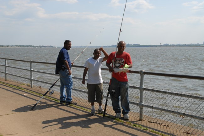 Three individuals fishing in East Potomac Park, two adults and one child. The man on the right is holding a catfish up for the camera to see.