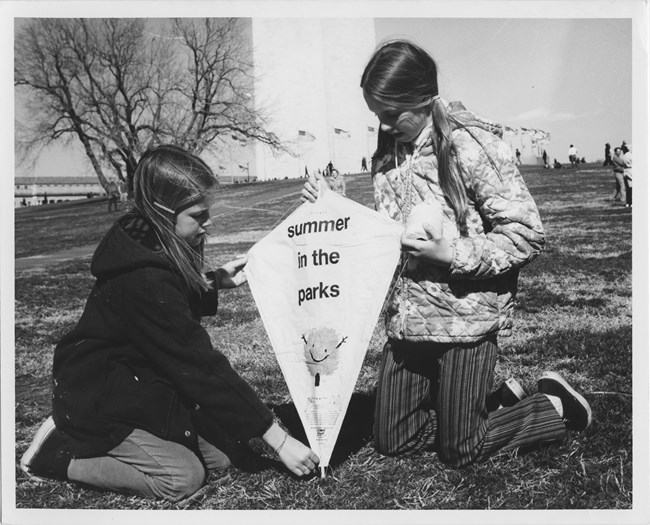 Two girls kneel with a kite that says "Summer in the parks" in front of the Washington Monument.