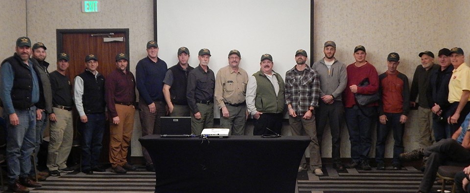 A group of 17 men stand in front of a room.