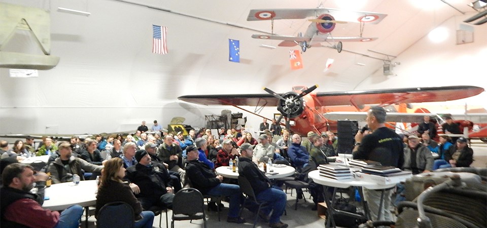A man with a microphone stands in front of a group in area displaying historic airplanes.