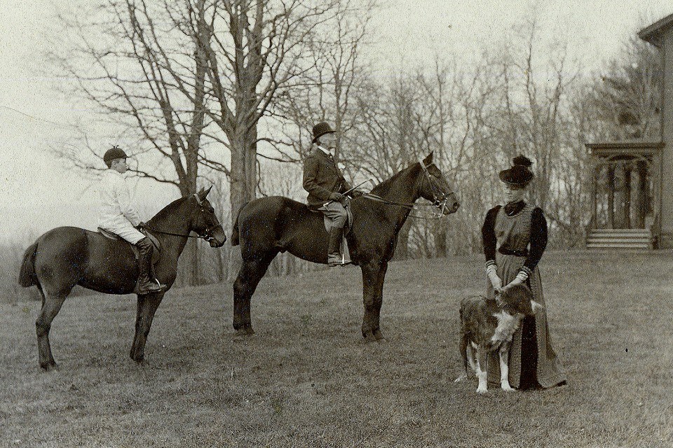 A boy and a man each on a horse. A woman with dog standing beside them. A house in the distance.