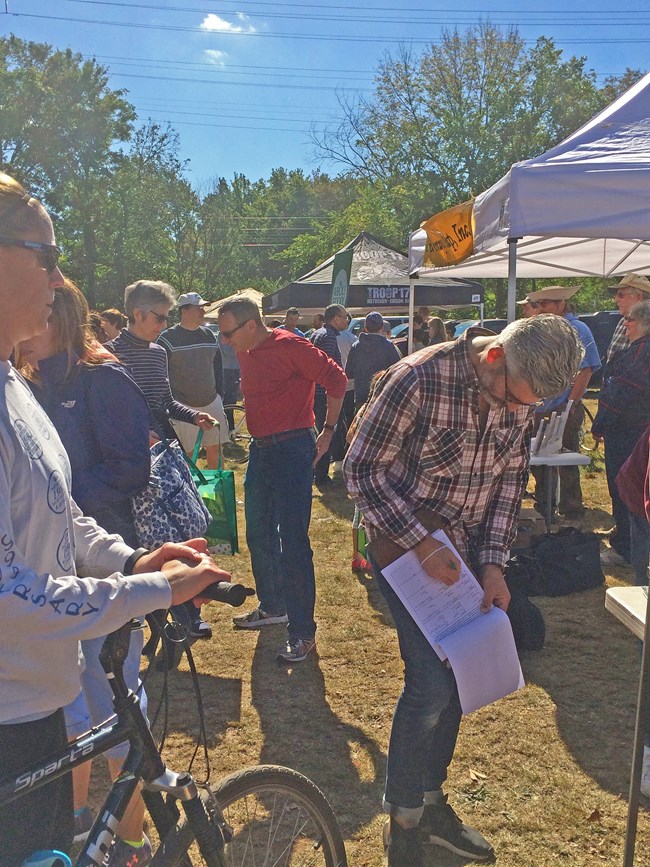 Jersey Residents Support People are outside next to pop-up tents. Man on a bike, man signing a clipboard and people talking.