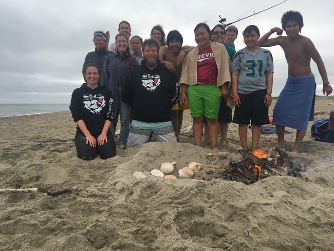 Teenagers stand on beach behind campfire and clay lamps.