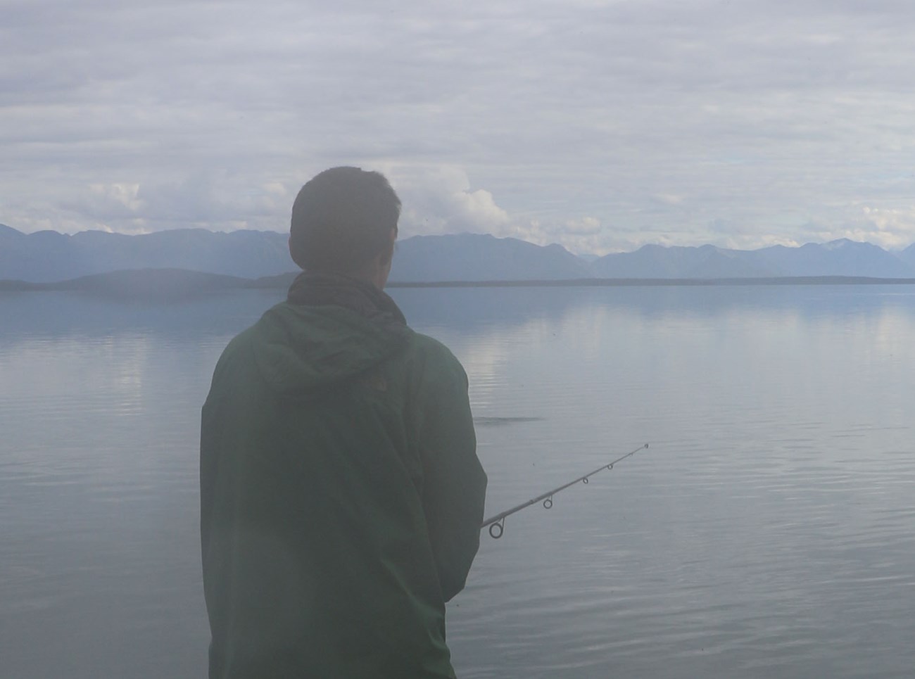 A young man stands on the shore of the Newhalen River in the fog while fishing with a rod and reel.