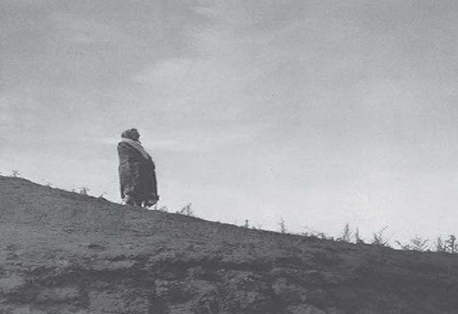 Adult covered in a blanket stands and faces east on a dirt mountain with sporadic shrubs.