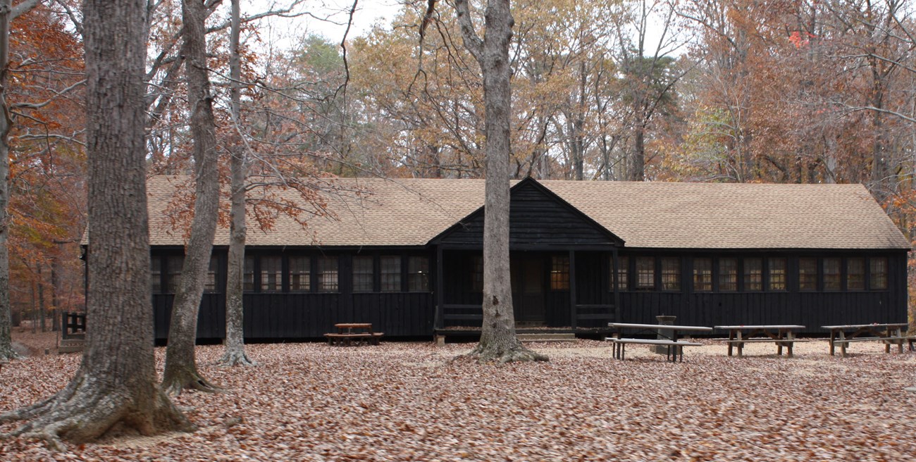 The Dining Hall is a long wooden Rustic style structure with a row of windows, under autumn trees.