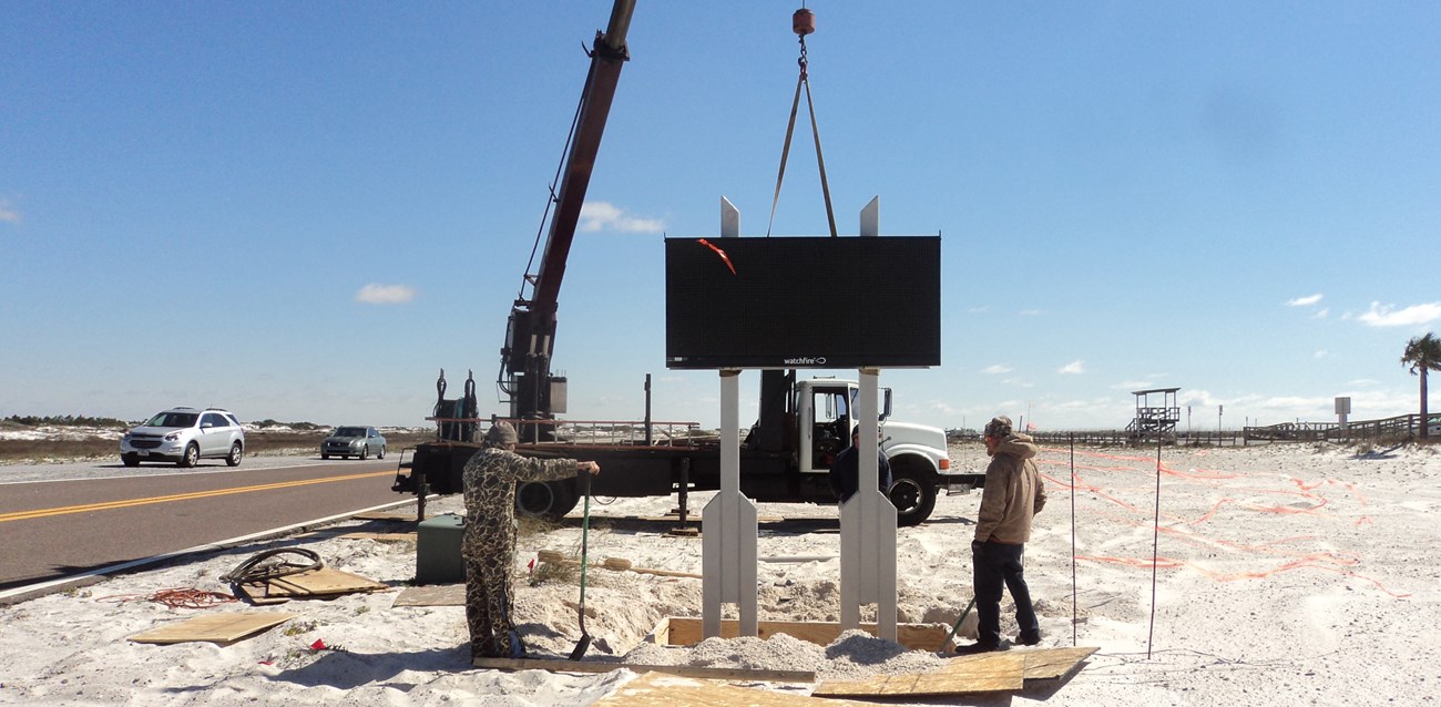 Crews install an electronic sign in sand along a road shoulder. Crews install an electronic sign in sand along a road shoulder.
