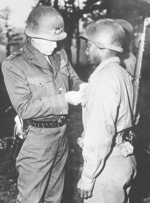 Two soldiers wearing helmets while one on left pins on an award onto soldier on right