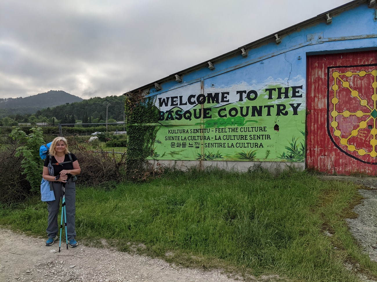 Deb Gardecki A person wearing a large backpack and carrying hiking polls stands near a building that has a mural painted on it that says Welcome to the Basque Country and in multiple languages reads Feel the Culture.