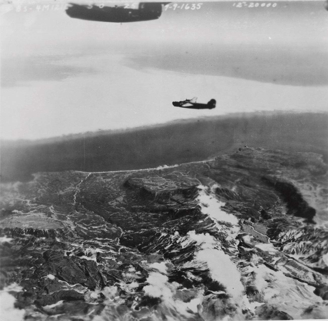 View from plane over a rugged coast line, with another plane in the sky.