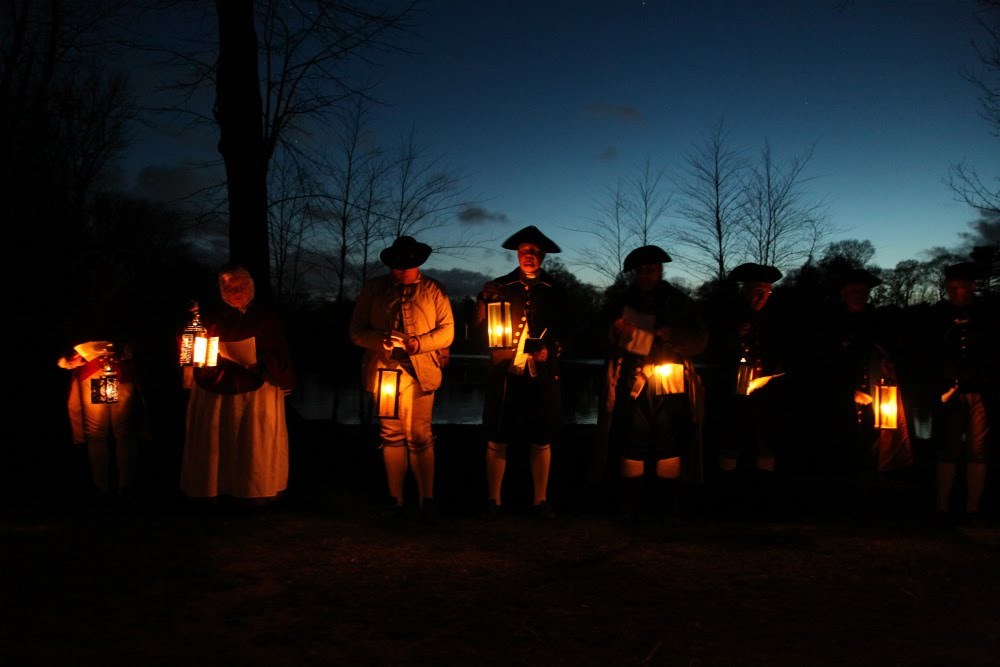 Men and women in period costumes hold lanterns at night.