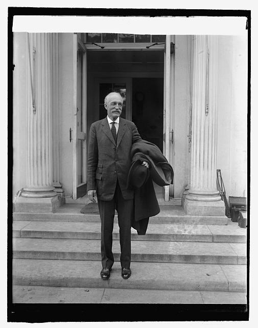 Black and white image of a man standing on steps outside of a building holding a jacket.