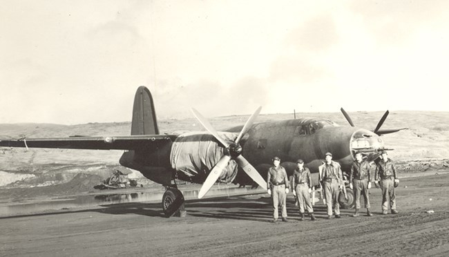 Group of men standing in front of plane, which has tarps over the engines.