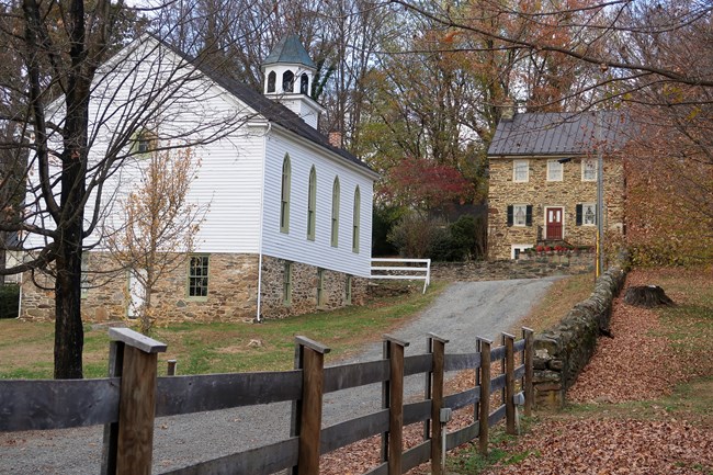 Color photo of a lane connecting Main Street and Bond Street runs past the John Wesley Church, the white wooden structure on the left, and the Samuel Means House, a stone house with 5 windows and a red door showing.