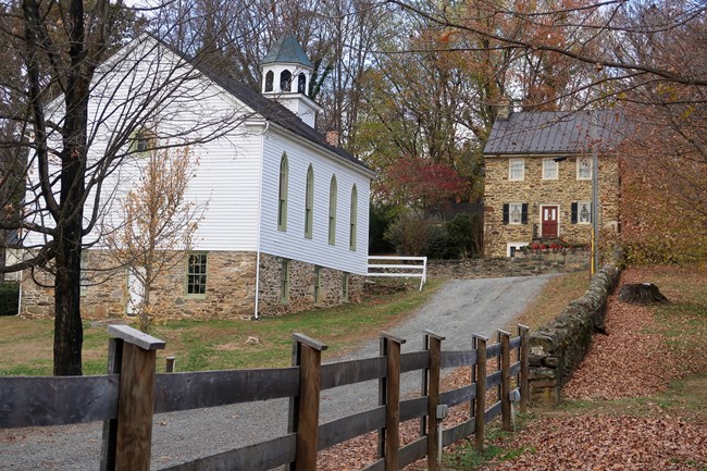 John Wesley Church and the Samuel Means House Color photo of a lane connecting Main Street and Bond Street runs past the John Wesley Church, the white wooden structure on the left, and the Samuel Means House, a stone house with 5 windows and a red door showing.
