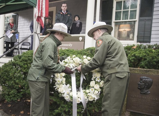 Two rangers are placing a flower wreath in front of the John Fitzgerald Kennedy National Historic Site home.