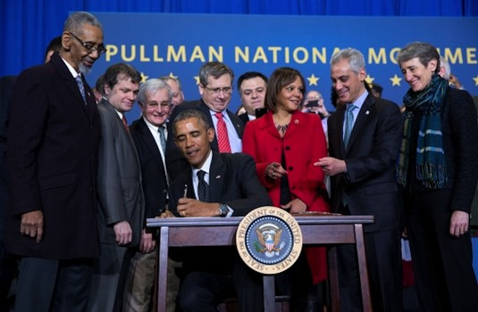 President Obama signing the establishment of Pullman National Monument. A group of about 10 people standing around President Obama sitting at a desk.