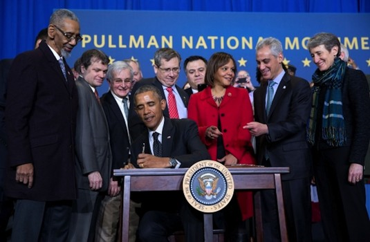 A group of about 10 people standing around President Obama sitting at a desk.