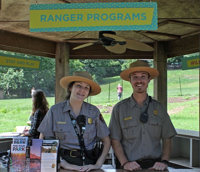 Two NPS park rangers pose in a wooden kiosk under a sign that says "Ranger Programs."