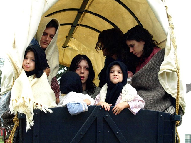 Seven people look forlorn in the back of a covered wagon.