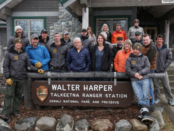 A smiling group of men and women stand behind a sign in front of a ranger station.