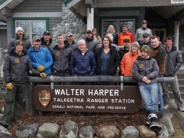 Talkeetna-based staff and contracted helicopter crew A smiling group of men and women stand behind a sign in front of a ranger station.