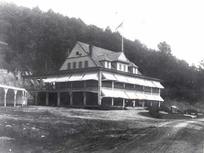 Black and white photo of the Rock Spring Club. The building is 3 stories, wrap around porch on the base floor, wrap around blacony on the second floor, and enclosed third floor with 9 windows showing. The first and second floor have light colored awnings.