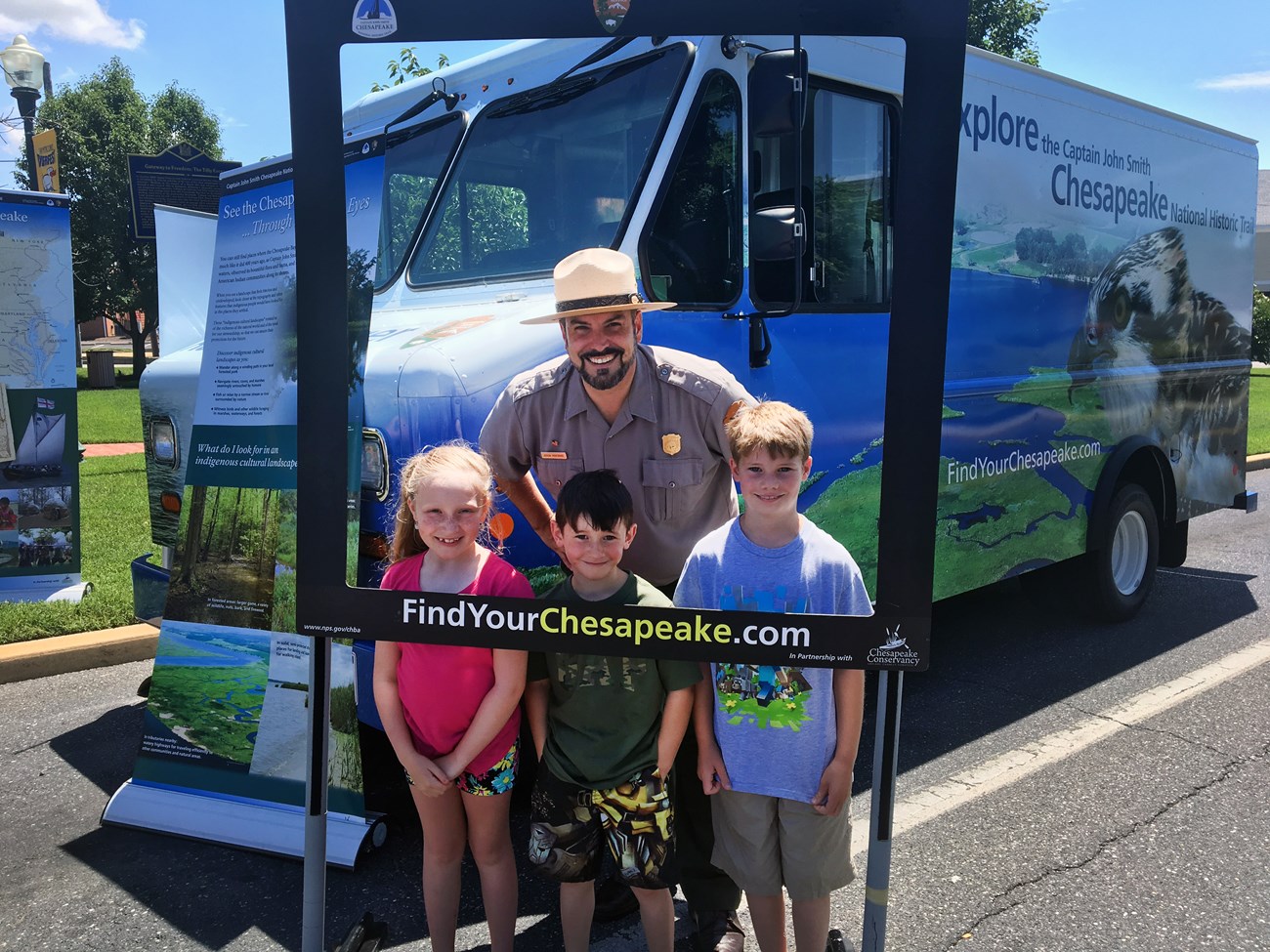 A male ranger poses in a "Selfie" frame with young kids. Mobile van is in the background.