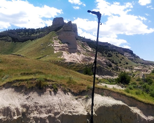 Bat acoustic recording equipment on a tall tripod in a grassy area with a bluff in the background