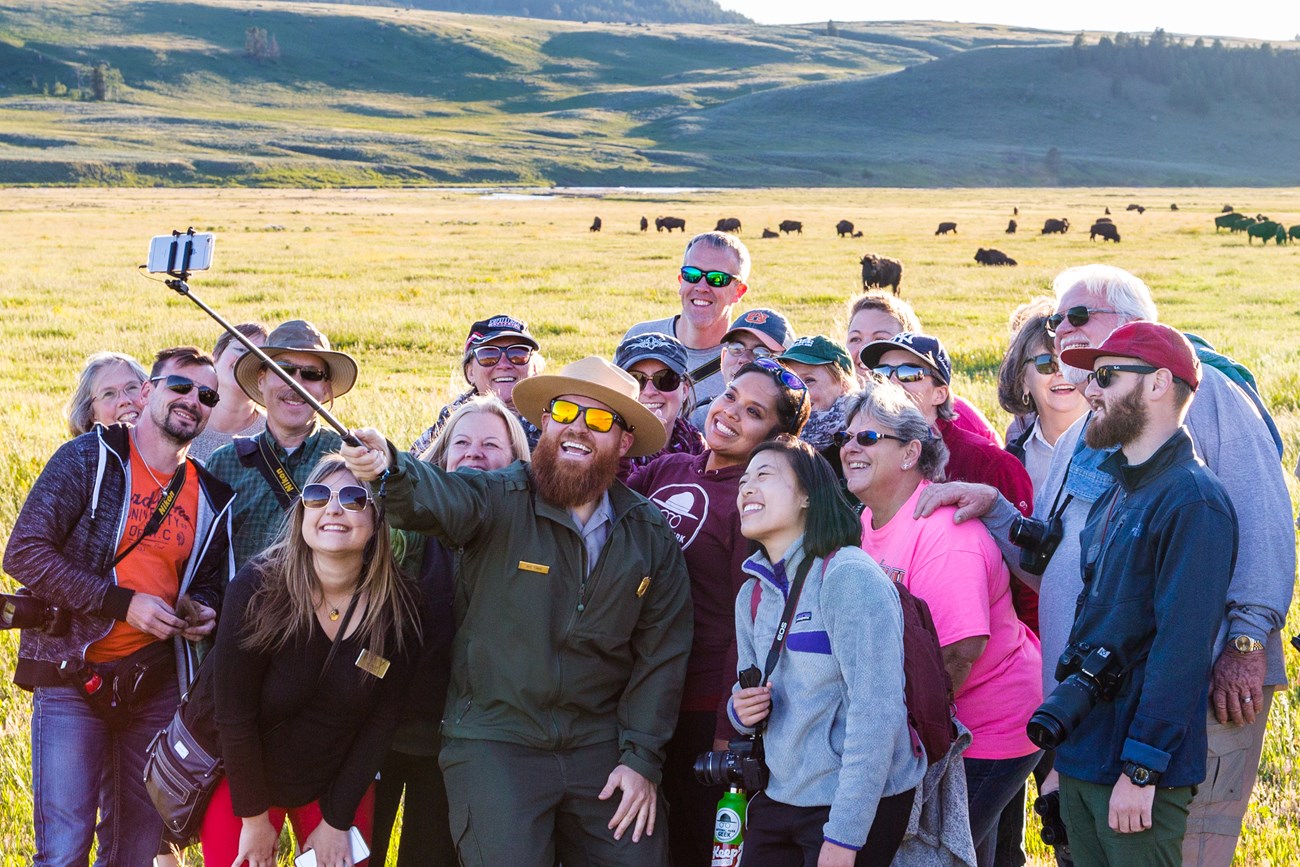 A park ranger and a group of visitors take a safe selfie at Yellowstone.  Bison are seen in the background at least 25 yards away.