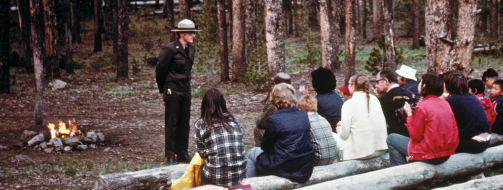 Paul Schullery delivering an evening program. A young man standing in front of an audience with a campfire in the background of a campground.