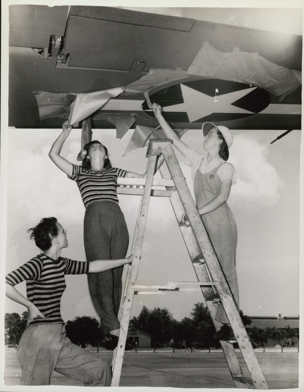 Women in defense work and in service (WAAC) black and white photo of three white women, two standing on a ladder, working on a ceiling