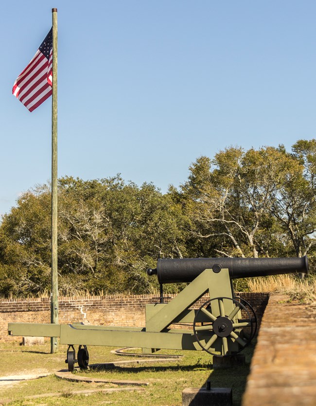 A cannon is mounted over a brick wall to the right, to the left an American flag flies from a wooden pole.