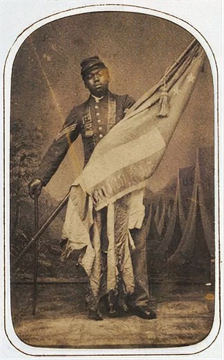 African American man in Civil War Union Army uniform holding an American flag leaning on a walking stick with tents in the background