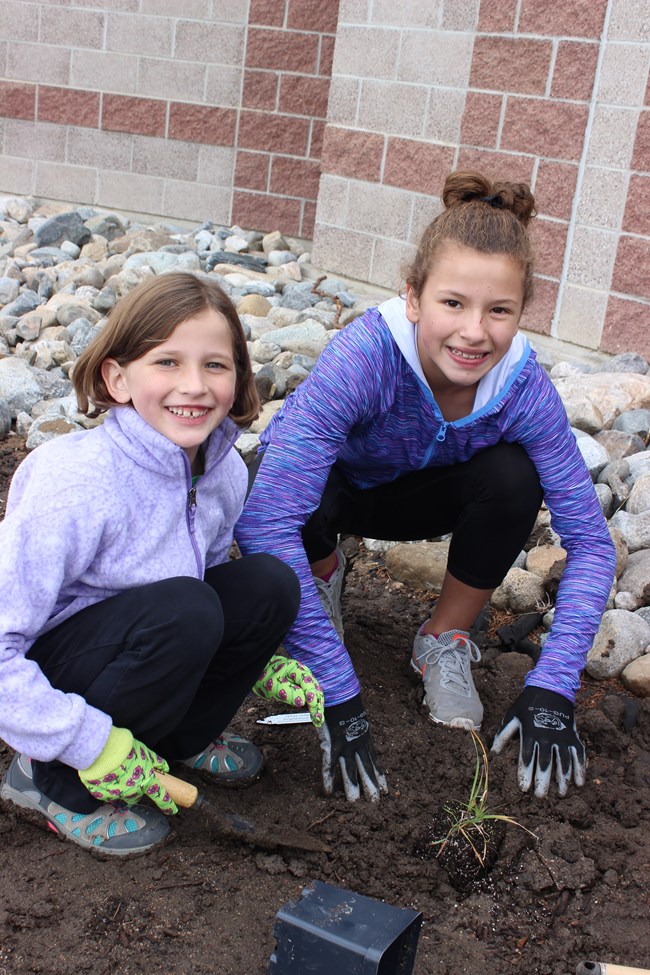 Two young girls smile at the camera as they place a small green plant in the ground