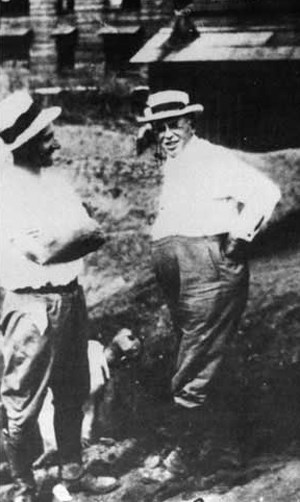 Two men standing in front of a building wearing fedora hats and long pants