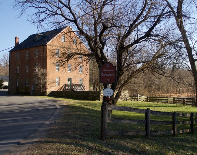 Color photo of a brick building, on the left, and a road sign that reads "Welcome to Waterford" in front of a large tree with no leaves