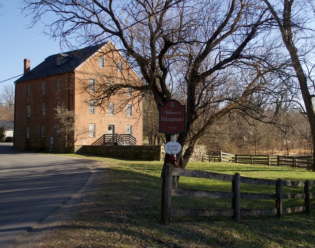 Waterford Mill Color photo of a brick building, on the left, and a road sign that reads "Welcome to Waterford" in front of a large tree with no leaves