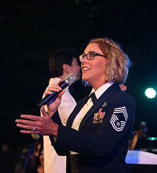 A woman with short curly blonde hair, and glasses, wearing a blue United States Air Force Dress uniform sings at an outdoor amphitheater at night.
